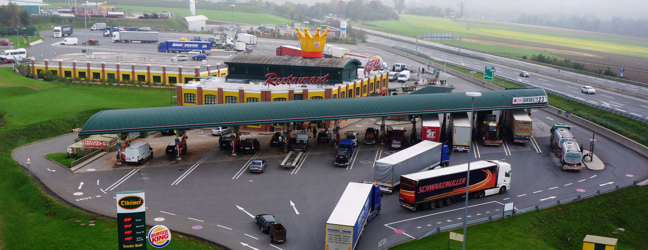 Außenansicht einer modernen Tankstelle mit angrenzender Raststation, umgeben von klar strukturierten Grünflächen und der Autobahn.