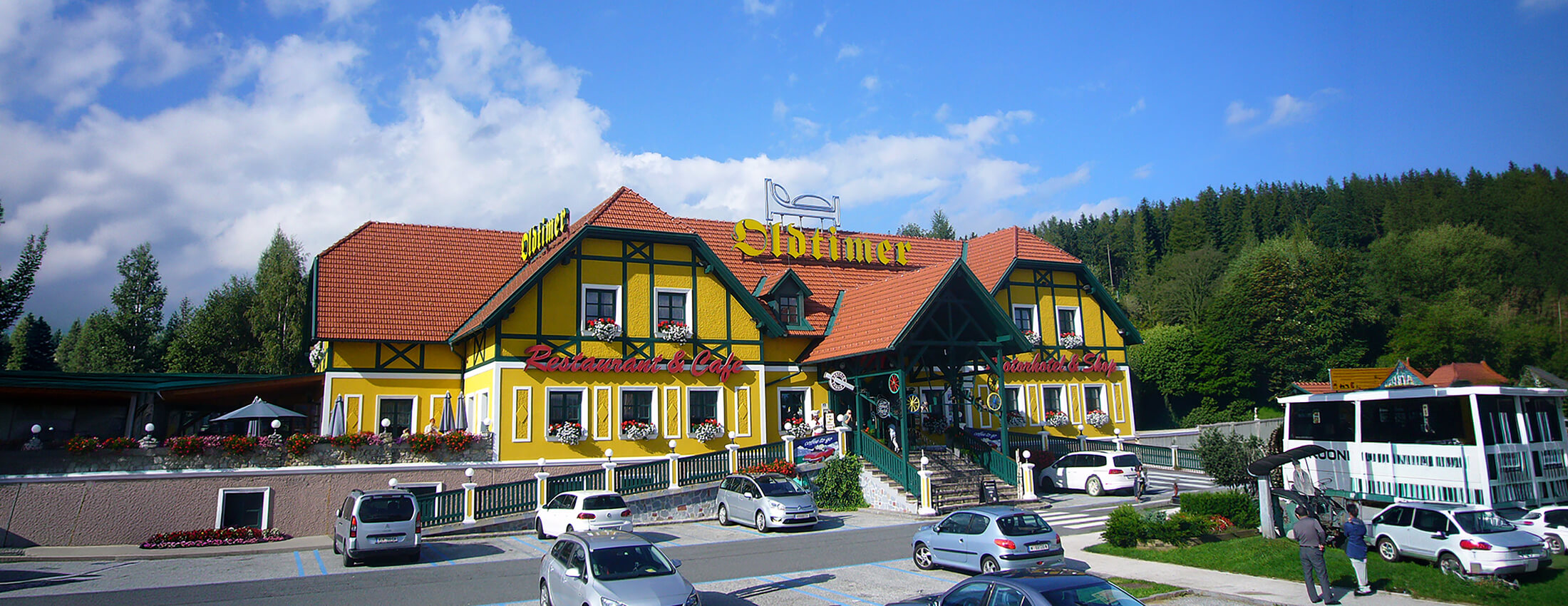 Blick auf den Oldtimer Standort Pack an einem sonnigen Tag. Parkende Autos und ein Bus befinden sich auf dem Parkplatz im Vordergrund.