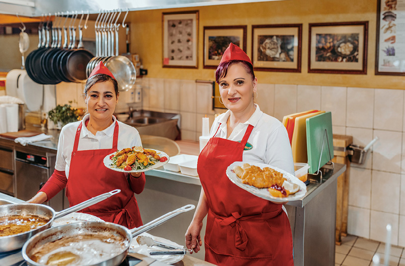Blick auf zwei Mitarbeiterinnen in der Küche, die fertige Gerichte präsentieren.